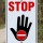 Das Bild zeigt ein Verkehrsschild mit dem Wort STOP in großen roten Buchstaben über einem schwarzen Handzeichen, das signalisiert, dass man anhalten soll. Dieses Schild wird häufig an Straßen oder Einfahrten verwendet, um Verkehrsteilnehmer auf eine Stoppsituation hinzuweisen.
