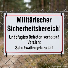 Das Bild zeigt ein Schild, das an einem Zaun in einem militärischen Sicherheitsbereich angebracht ist. Es warnt vor unbefugtem Betreten und weist darauf hin, dass Schusswaffengebrauch droht.