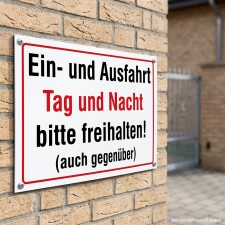 Das Bild zeigt ein Schild, das an einer Wand angebracht ist und darauf hinweist, dass die Ein- und Ausfahrt sowohl tagsüber als auch nachts freigehalten werden soll. Der Text ist in einer Kombination aus schwarzer und roter Schrift auf weißem Hintergrund gestaltet, um die Aufforderung deutlich zu machen.