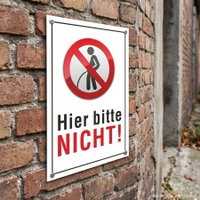 Das Bild zeigt ein Schild, das an einer Backsteinmauer befestigt ist. Es trägt die Aufschrift Hier bitte NICHT! in großen, roten Buchstaben und enthält ein Symbol, das eine verbotene Handlung darstellt.