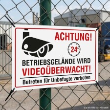 Das Bild zeigt ein Warnschild, das an einem Zaun angebracht ist und darauf hinweist, dass das Betriebsgebiet videoüberwacht wird. Es enthält die Aufschrift ACHTUNG! sowie Informationen zur 24-Stunden-Überwachung und den Hinweis, dass das Betreten für Unbefugte verboten ist.