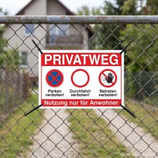 Das Bild zeigt ein Schild mit dem Text PRIVATWEG, das an einem Zaun befestigt ist. Es weist darauf hin, dass Parken, Durchfahren und Betreten des Weges verboten sind und ist für die Nutzung nur durch Anwohner gedacht.