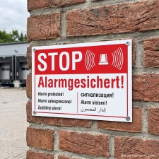 Das Bild zeigt ein rotes Schild mit der Aufschrift STOP Alarmgesichert! in großen, auffälligen Buchstaben. Es ist an einer Ziegelwand angebracht und informiert darüber, dass der Bereich durch ein Alarmsystem gesichert ist, um unbefugten Zutritt zu verhindern.