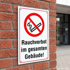 Das Bild zeigt ein Schild mit einem roten Durchmesser-Symbol, das eine durchgestrichene Zigarette darstellt, und dem Text „Rauchverbot im gesamten Gebäude!“ in großen, schwarzen Buchstaben. Dieses Schild ist an einer Ziegelwand angebracht und weist darauf hin, dass das Rauchen in dem gesamten Gebäude nicht erlaubt ist.
