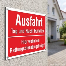 Das Bild zeigt ein rotes Schild mit der Aufschrift „Ausfahrt Tag und Nacht freihalten!“ und darunter „Hier wohnt ein Rettungsdienstangehöriger!“. Das Schild ist an einer Wand neben einem Eingangsbereich angebracht und dient dazu, darauf hinzuweisen, dass die Ausfahrt jederzeit frei bleiben muss, um den Zugang für Rettungsdienste zu gewährleisten.