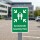 Das Bild zeigt ein grünes Schild mit der Aufschrift Sammelstelle Assembly Point. Es ist in einem Parkplatz aufgestellt und weist in alle vier Himmelsrichtungen auf einen Sammelplatz für Personen hin, was auf einen Sicherheitskontext hinweist.