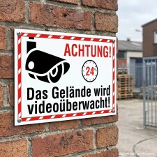 Das Bild zeigt ein Warnschild, das an einer Backsteinmauer befestigt ist. Es informiert mit dem Text „Achtung! Das Gelände wird videoüberwacht!“ und ist mit einem Symbol einer Überwachungskamera sowie einem Hinweis auf die 24-Stunden-Überwachung versehen, um Passanten auf die Sicherheitsmaßnahmen hinzuweisen.