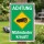 Das Bild zeigt ein Warnschild mit dem Text ACHTUNG Mähroboter kreuzt! auf einem grünen Hintergrund. Das Schild ist in einem Garten aufgestellt und weist darauf hin, dass ein Mähroboter in der Nähe unterwegs ist, um Passanten zu warnen.