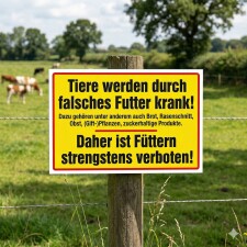 Das Bild zeigt ein auffälliges Schild, das auf einem Holzpfosten in einem Feld angebracht ist. Es warnt davor, dass Tiere durch falsches Futter, wie Brot und Obst, krank werden können, und erklärt, dass das Füttern strengstens verboten ist.