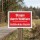 Das Bild zeigt ein rotes Warnschild, das auf eine Gefährdung durch Wildtiere hinweist, mit der Aufschrift „Staupe durch Wildtiere“ und dem Zusatz „Gefährdeter Bezirk“. Es steht an einem Weg in einem ländlichen Gebiet, um Passanten vor möglichen Risiken zu warnen.
