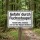 Das Bild zeigt ein weißes Schild, das auf einer Holzpfahl steht und im Wald platziert ist. Es warnt vor der Gefahr durch Fuchsstaupe und weist darauf hin, dass Hunde an der Leine gehalten und auf den Wegen bleiben sollen.