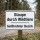 Das Bild zeigt ein Schild mit dem Hinweis „Staupe durch Wildtiere“ und dem Zusatz „Gefährdeter Bezirk“. Es befindet sich in einem Waldgebiet und warnt Passanten vor der möglichen Gefahr von Wildtieren, die an Staupe erkrankt sein könnten.
