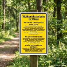 Das Bild zeigt ein gelbes Schild mit wichtigen Informationen zur Staupe, das in einem Waldgebiet aufgestellt ist. Es informiert über die Gefahren des Staupe-Virus für Hunde und gibt Hinweise zur Vorbeugung, insbesondere zur Impfung.