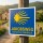 Das Bild zeigt ein Schild des Jakobswegs, das auf einem Holzpfosten montiert ist. Es hat einen blauen Hintergrund mit gelben Schriftzügen und einem Sonnenmotiv, und es dient als Wegweiser für Pilger auf dem Weg nach Santiago de Compostela.