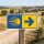 Das Bild zeigt ein Wegweiser-Schild für den Jakobsweg, das auf der linken Seite ein gelbes Symbol mit einer stilisierten Sonne und Strahlen zeigt, während die rechte Seite einen blauen Pfeil nach rechts anzeigt. Das Schild steht an einem Weg in einer ländlichen Umgebung und dient Pilgern als Orientierungshilfe auf ihrem Weg.