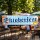Das Bild zeigt ein großes, farbenfrohes Banner mit der Aufschrift Oktoberfest, das in einem Biergarten aufgehängt ist. Es ist umgeben von einer blauen und weißen Rautenstruktur, die typisch für bayerische Motive ist, und zeigt neben der Schrift eine Brezel und ein Bierglas, was auf das Fest und die bayerische Kultur hinweist.