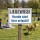 Das Bild zeigt ein Schild mit der Aufschrift LIEGEWIESE und darunter Hunde sind hier erlaubt! in blauer Schrift auf weißem Hintergrund. Das Schild steht in einem Park, wo Menschen auf einer Wiese sitzen und Hunde umherlaufen.