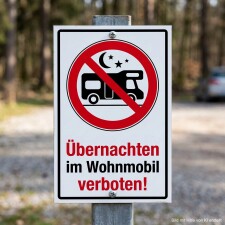 Das Bild zeigt ein Schild, das deutlich darauf hinweist, dass das Übernachten im Wohnmobil verboten ist. Es ist in einem Waldgebiet aufgestellt und enthält ein Symbol eines Wohnmobils mit einem roten Durchgestrichenen, um die Regelung zu verdeutlichen.